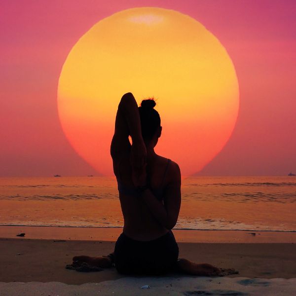 Silhouette of a person meditating in a lotus position on the beach at sunset with a colorful sky.