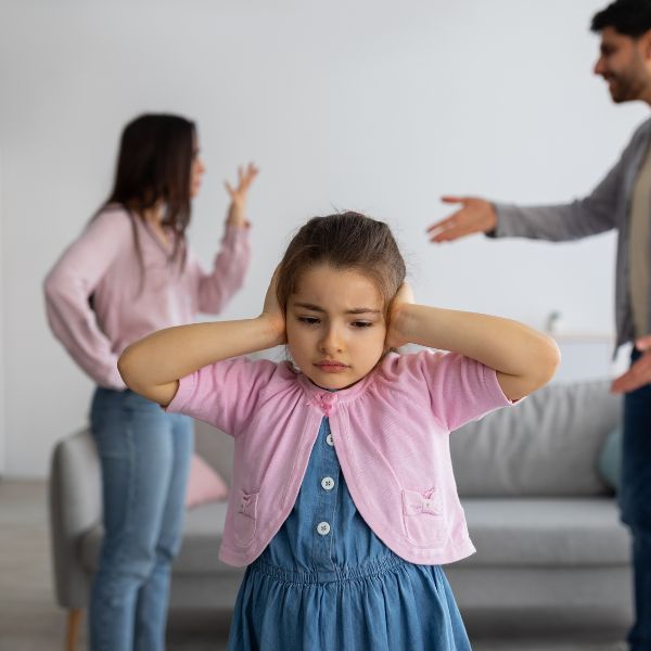 A young girl covers her ears while her parents argue in the background.