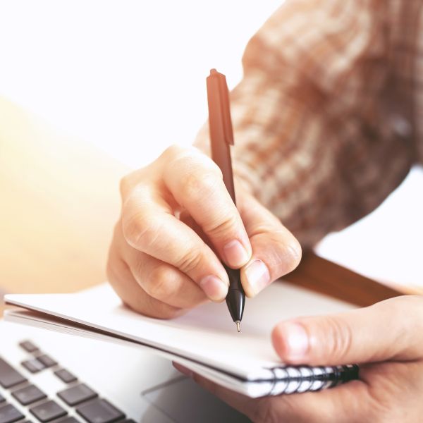 Person writing in a notebook beside a laptop, representing preparation and planning for healing sessions.